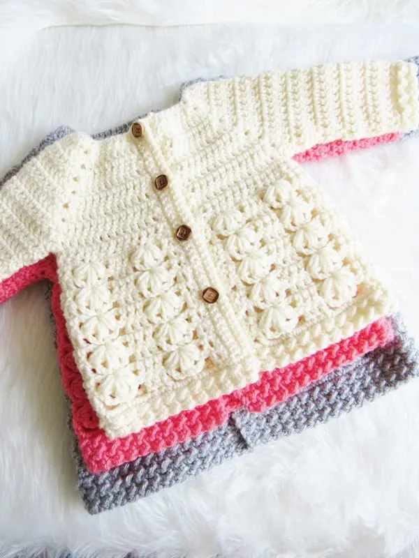 Crochet baby sweaters in 3 sizes piled on top of each other, gray, pink and cream on top. 