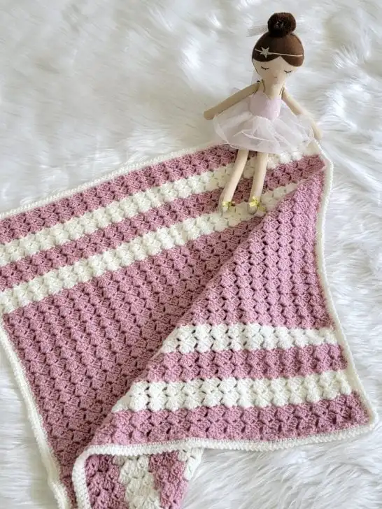Crochet blanket in pink and cream color with a ballerina doll sitting on it
