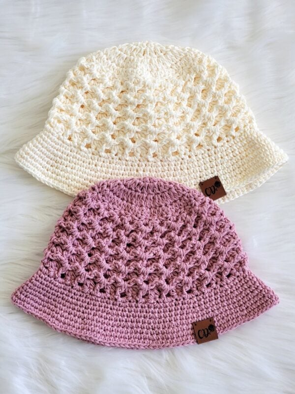 Two handmade crochet bucket hats in cream and dusty pink displayed on a soft white surface, featuring a textured open stitch crown and solid brim.