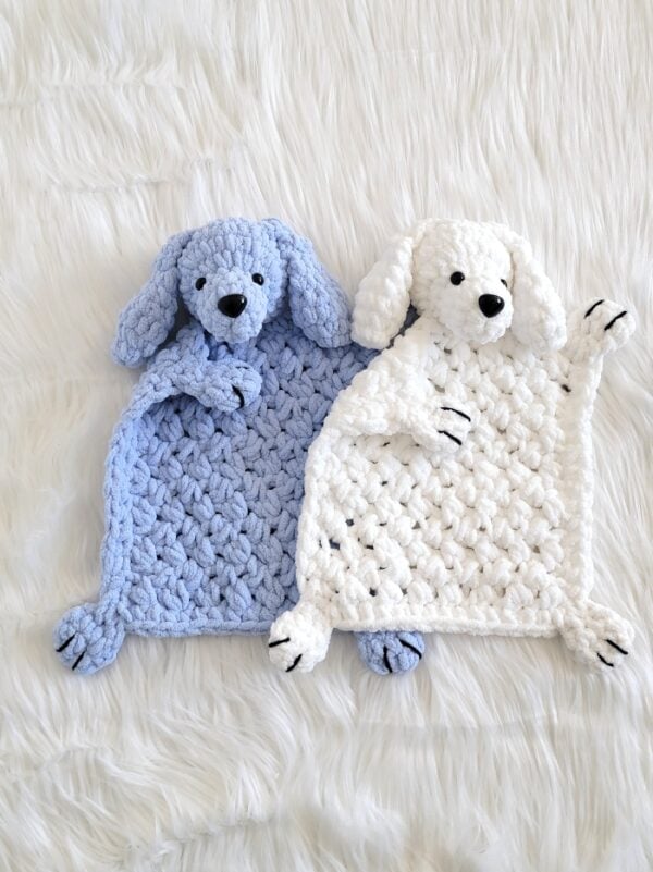 Two plush crochet dog loveys in pastel blue and white laid side by side on a soft white faux fur blanket.