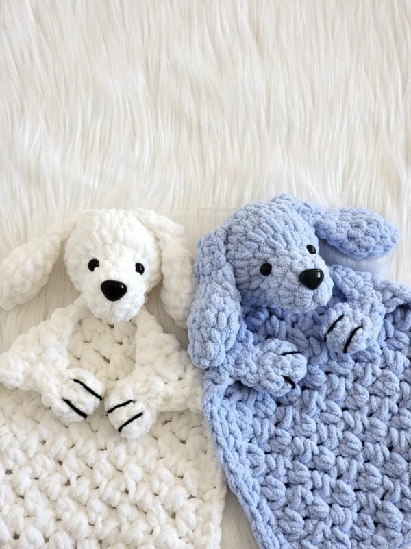 White and pastel blue crochet dog loveys with floppy ears and chunky textured stitching resting on a white faux fur background.