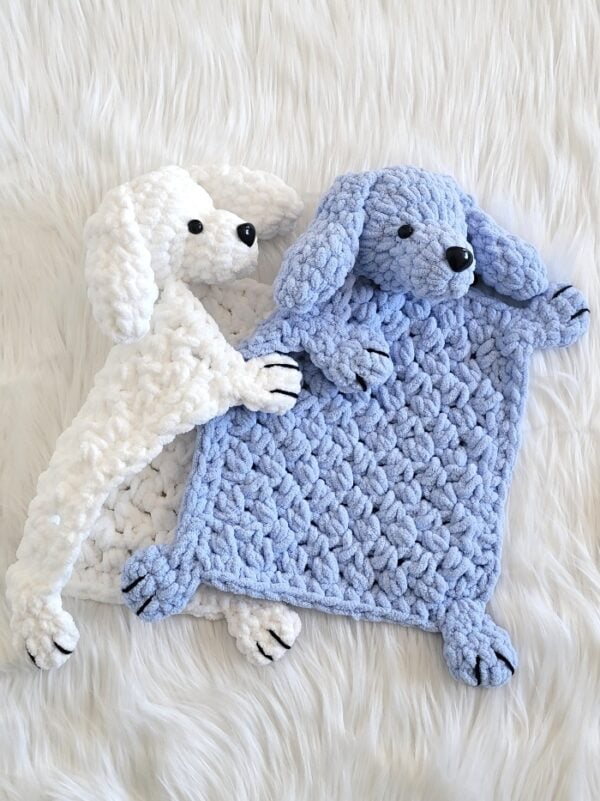 White crochet dog lovey with floppy ears hugging blue lovey and resting on a white faux fur background.