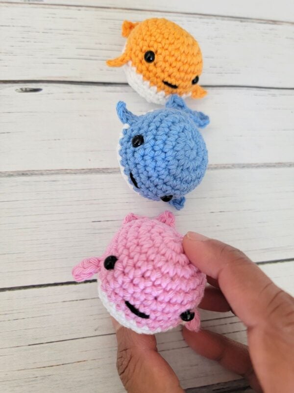 Three tiny crochet whale plushies in pastel pink, blue, and orange.