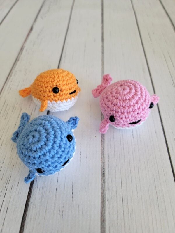 Tiny crochet whales in pink, blue, and orange on a white wooden background.