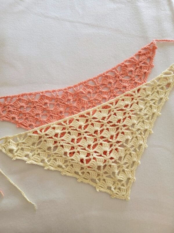 Two handmade crochet bandanas in cream and peach cotton yarn featuring a delicate shell stitch lace pattern, displayed overlapping on a white fabric background with long tie straps visible.