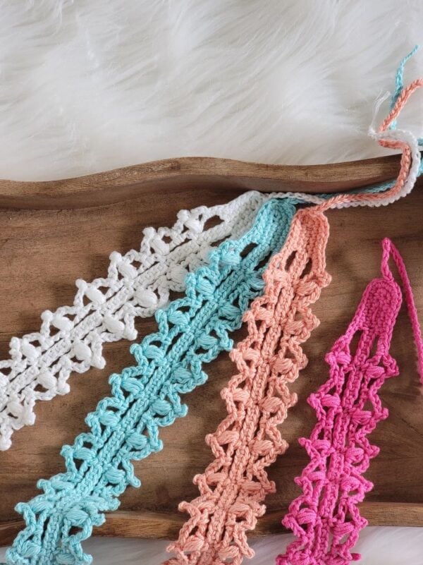 Four lacy crochet headbands in white, aqua, coral, and pink displayed on a wooden tray.