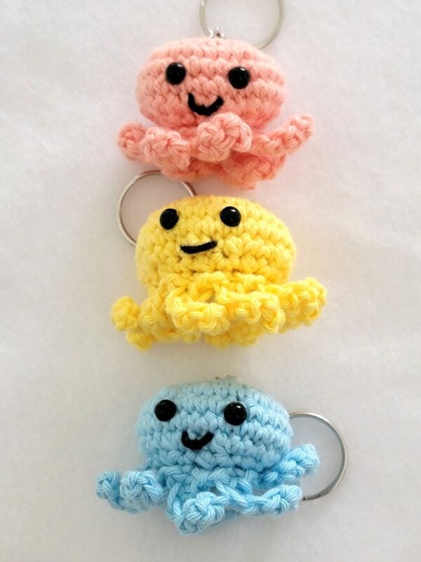Pink, yellow, and blue crochet octopus keychains arranged vertically on a white background.