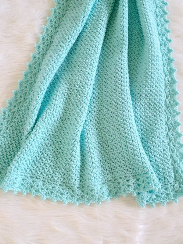 Aqua crochet baby blanket with a simple textured stitch and delicate lacy border, shown softly draped on white fur.