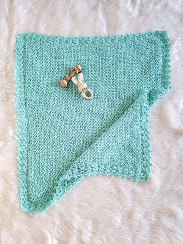 Simple aqua crochet baby blanket laid flat on white fur with one corner folded back and a wooden baby toy on top.