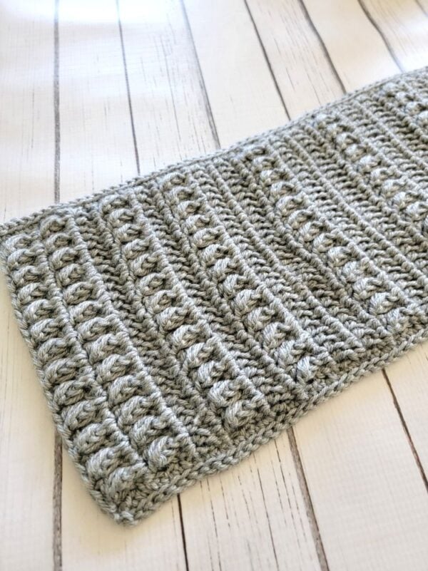 Gray men’s crochet scarf with raised textured stitch rows displayed flat on a white wood surface.