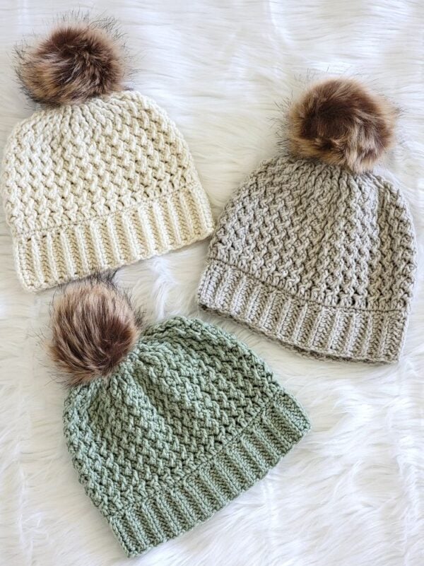 Three handmade crochet hats in cream, taupe, and sage green with ribbed brims and faux fur pom poms.