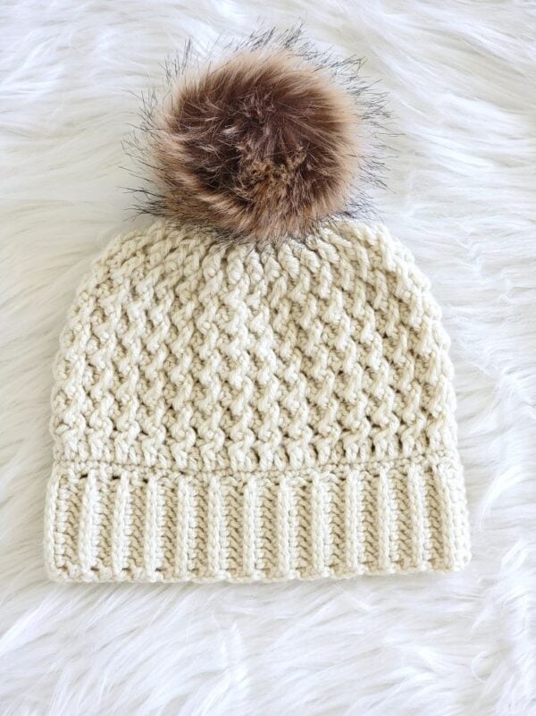 Cream crochet winter hat with a ribbed brim, textured stitches, and a brown faux fur pom pom.
