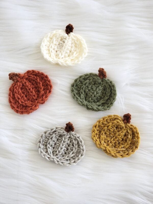 Five crochet pumpkin appliqués in cream, rust orange, olive green, gray, and mustard yellow on a white faux fur background.