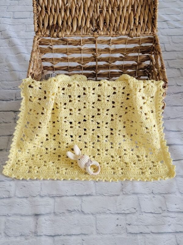 Soft yellow crochet baby blanket with an openwork flower design draped over a wicker basket with a bunny rattle.