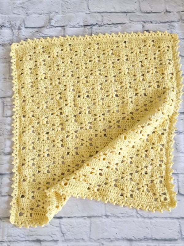 Pale yellow crochet baby blanket with raised flower stitches, a delicate border, and one corner folded over.