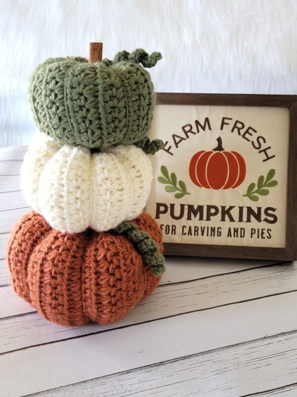 Three crochet pumpkins in sage green, cream, and orange stacked together beside a rustic fall pumpkin sign.