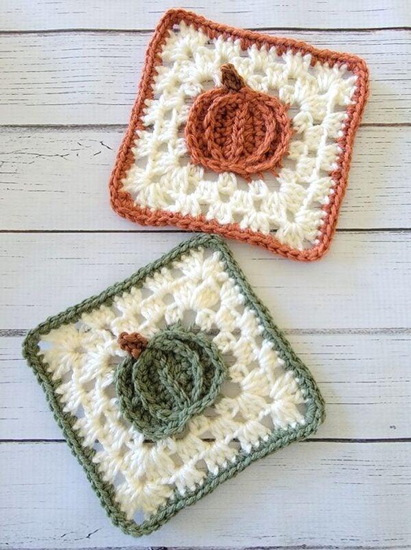 Two handmade crochet pumpkin coasters arranged diagonally on a white wooden background, featuring orange and green pumpkin granny squares.