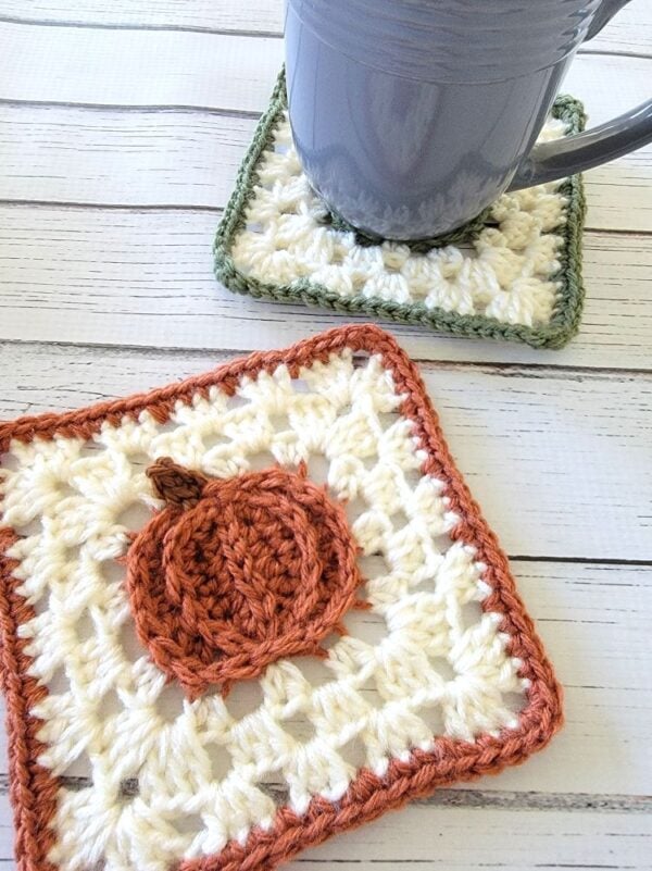 Handmade orange pumpkin crochet coaster in the foreground with a green pumpkin coaster holding a gray mug in the background.